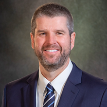 Professional headshot of a person in a suit with a dark, blurry background.
