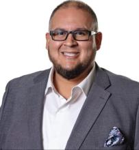 Professional headshot of a person wearing a gray suit and glasses, smiling.