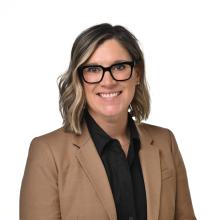 Professional headshot of a person wearing a tan blazer and black glasses, smiling against a white background.