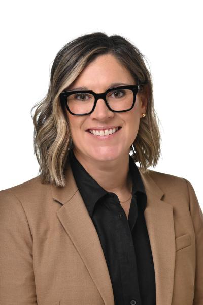 Professional headshot of a person wearing a tan blazer and black glasses, smiling against a white background.