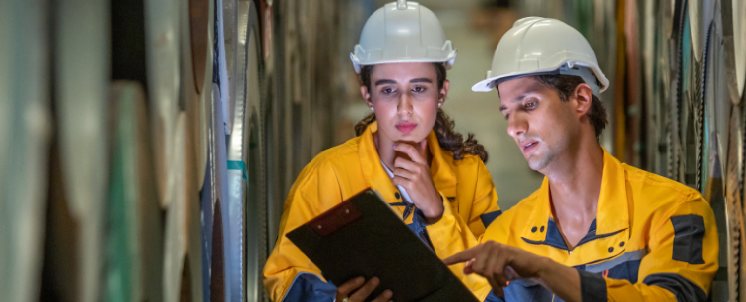 Two workers in safety helmets and industrial uniforms review a clipboard